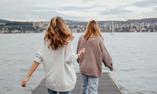 two women walk to the edge of a dock, in a body of water, looking toward the city on the other shore